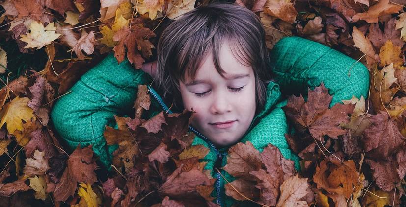 kleines Kind liegt mit geschlossenen Augen in einem bunten Laubhaufen