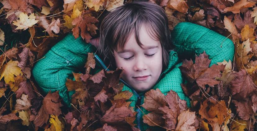 kleines Kind liegt mit geschlossenen Augen in einem bunten Laubhaufen
